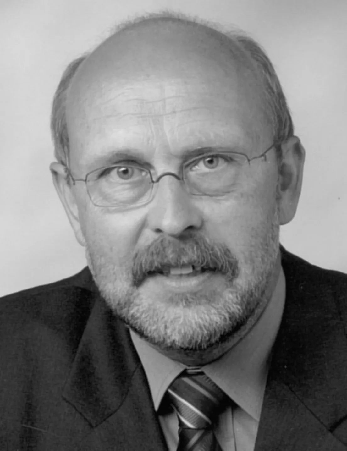 Porträt von Lothar Schütz, einem älteren Mann mit Brille, Bart und kurzem Haar, der einen Anzug und eine Krawatte trägt.