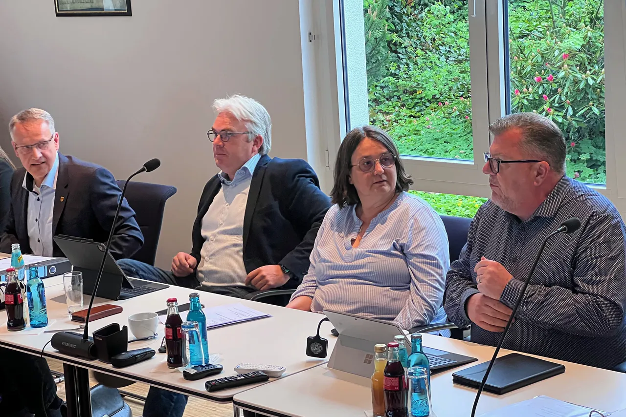Vier Personen sitzen an einem Konferenztisch und diskutieren. Ein Mann mit Brille spricht, während die anderen zuhören. Auf dem Tisch stehen Wasserflaschen, Mikrofone und Laptops.
