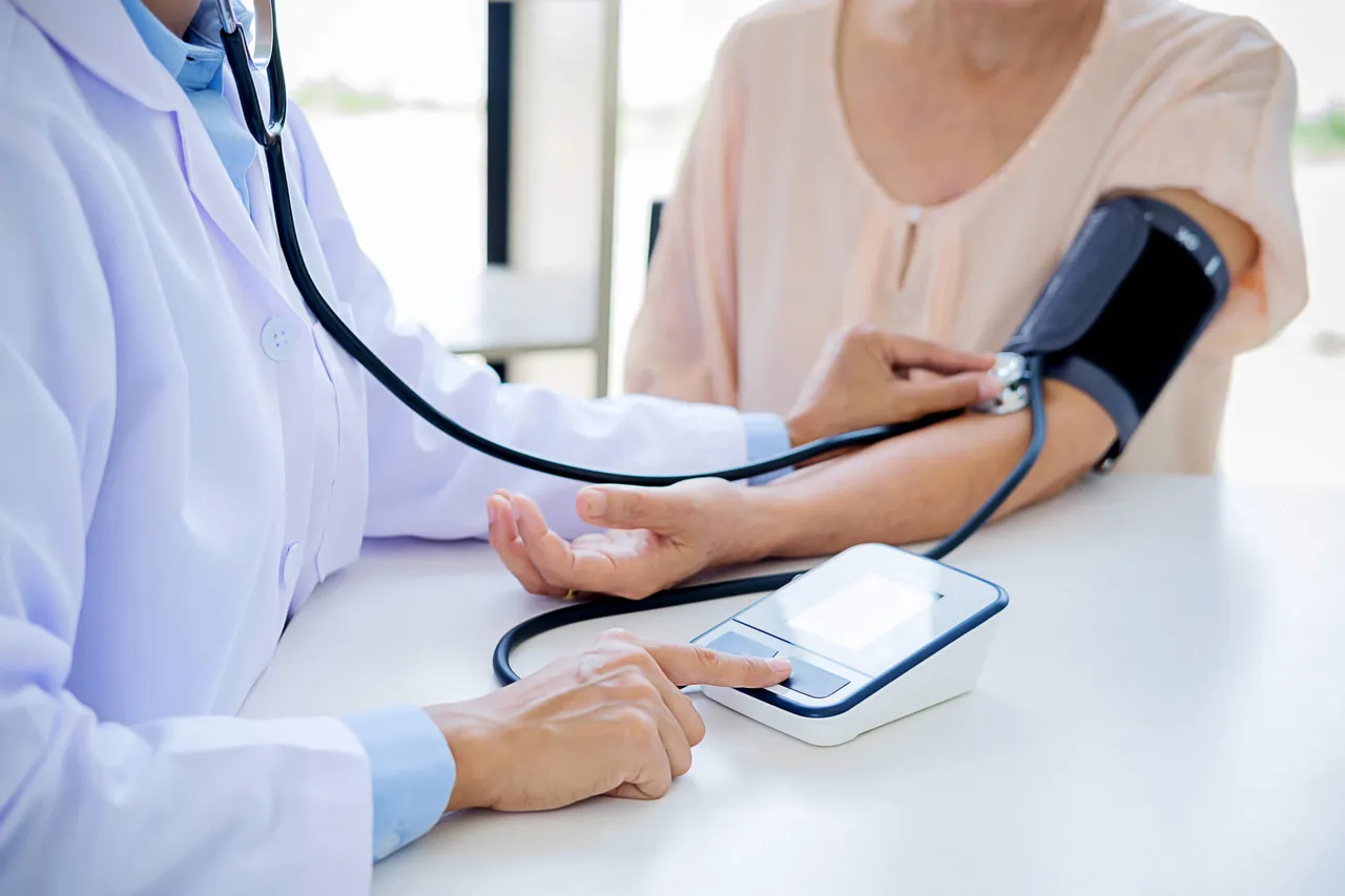 Ein Arzt misst einer Patientin den Blutdruck. Er bedient ein digitales Blutdruckmessgerät, während die Manschette am Arm der Patientin angelegt ist.