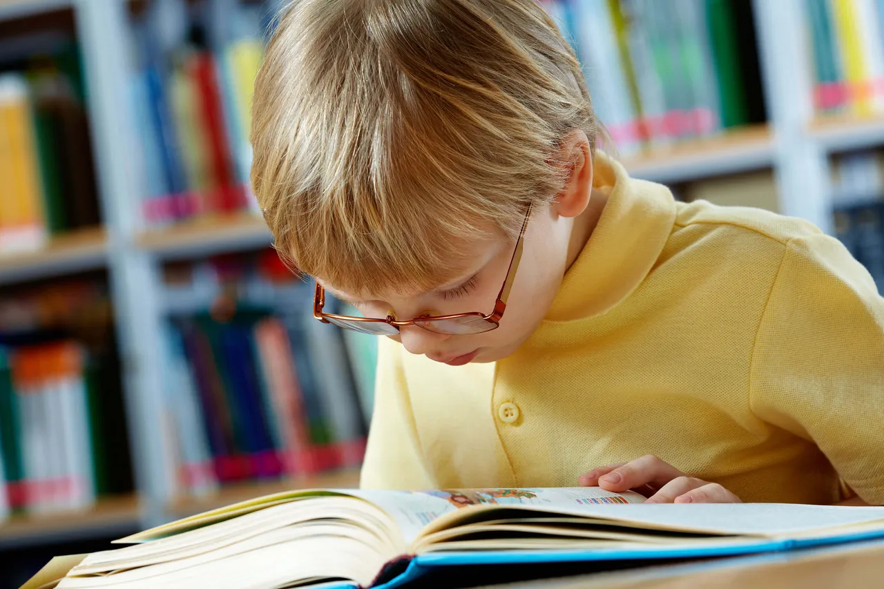 Ein Junge mit Brille liest konzentriert in einem aufgeschlagenen Buch vor einem Bücherregal.
