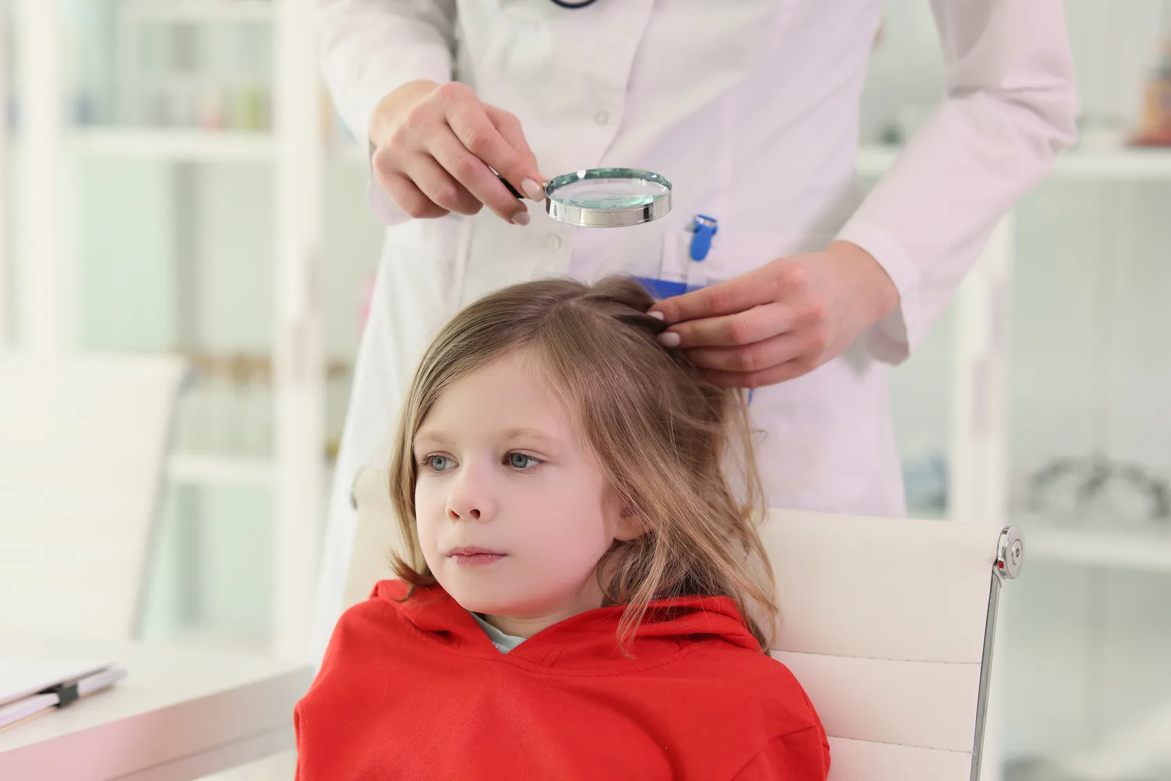 Eine Ärztin untersucht mit einer Lupe die Haare eines kleinen Mädchens. Das Mädchen trägt einen roten Pullover und sitzt auf einem Stuhl.