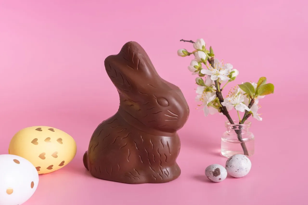 Schokoladenhase und Ostereier vor rosa Hintergrund. Eine blühende Kirschzweige steht in einem kleinen Glas daneben.