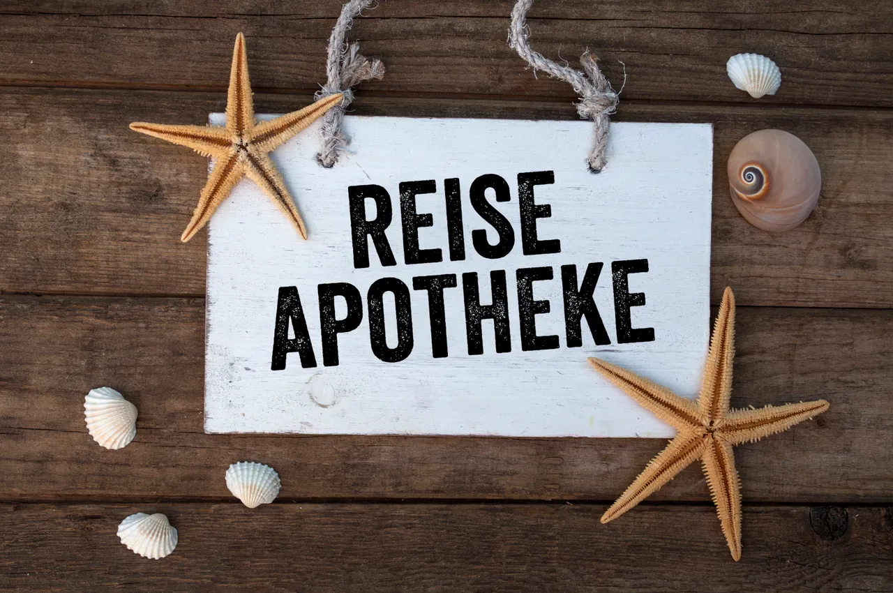 Ein weißes Holzschild mit der Aufschrift „REISE APOTHEKE“ liegt umgeben von Muscheln und Seesternen auf einem dunklen Holzbrett.