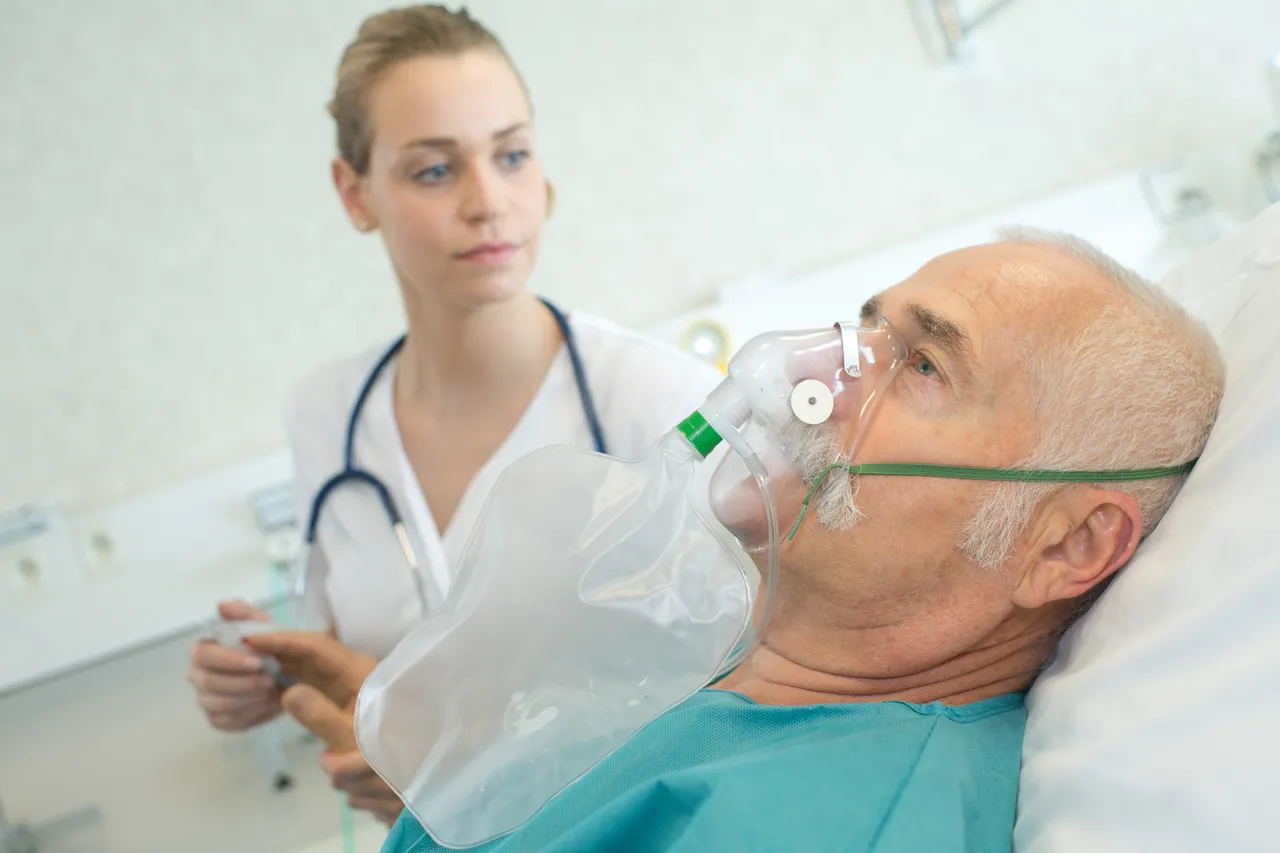 Ein älterer Mann im Krankenhausbett erhält Sauerstoff über eine Maske, während eine junge Ärztin im Hintergrund aufmerksam beobachtet.