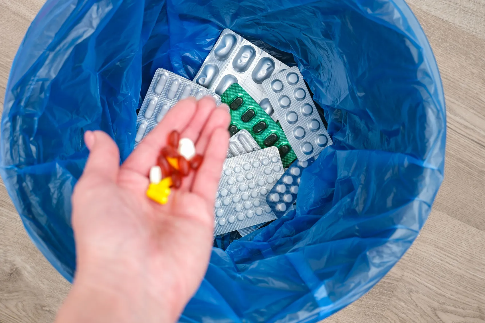 Hand wirft verschiedene Medikamente und leere Blisterpackungen in einen blauen Müllsack.
