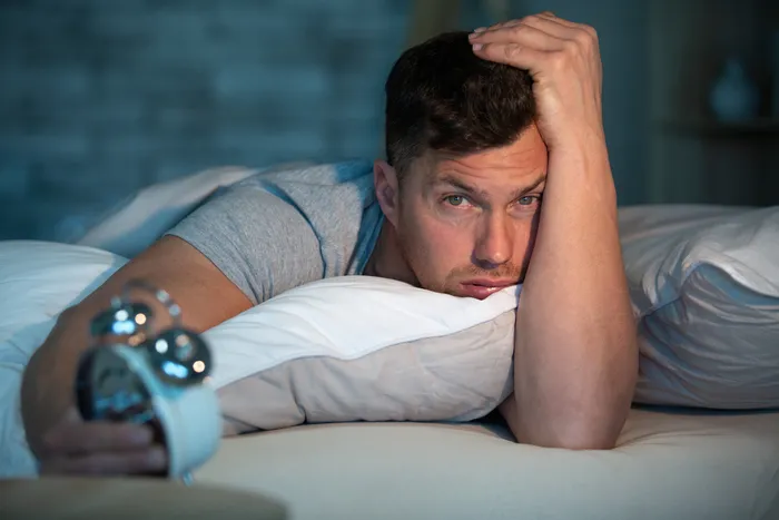 Ein Mann liegt wach im Bett und starrt besorgt in die Dunkelheit. Neben ihm steht ein Wecker, der auf kurz nach drei Uhr eingestellt ist.