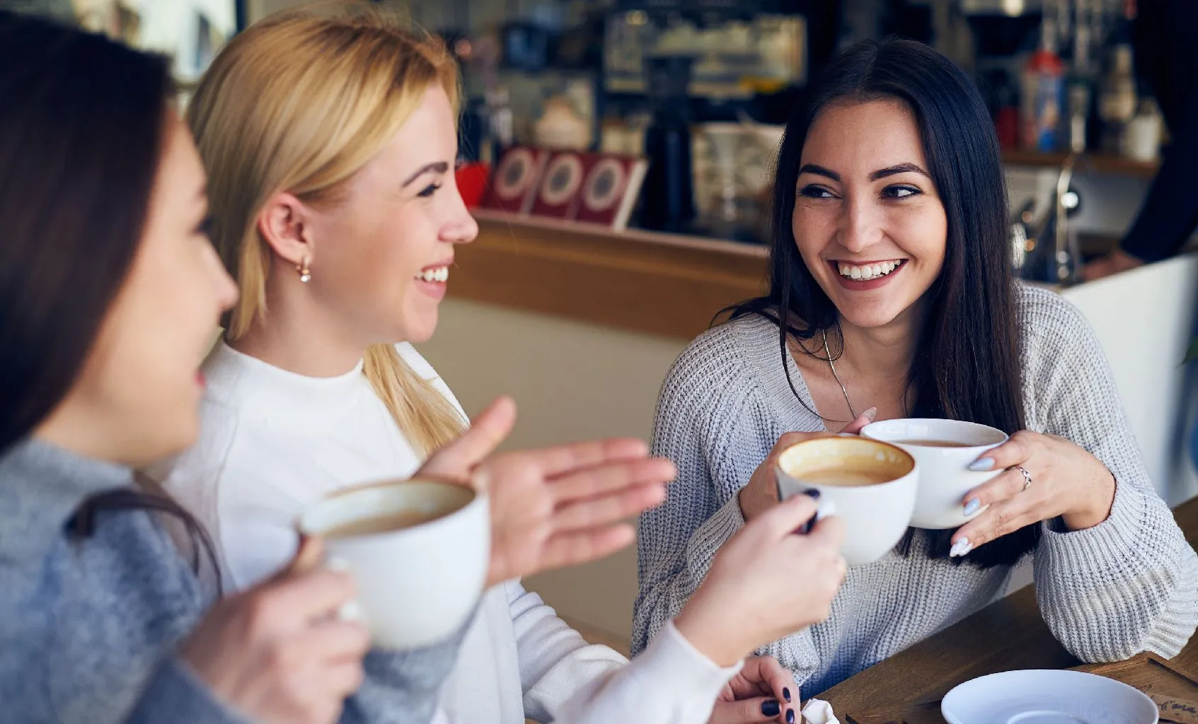 Drei junge Frauen sitzen lächelnd in einem Café zusammen und halten Tassen mit Kaffee in den Händen.
