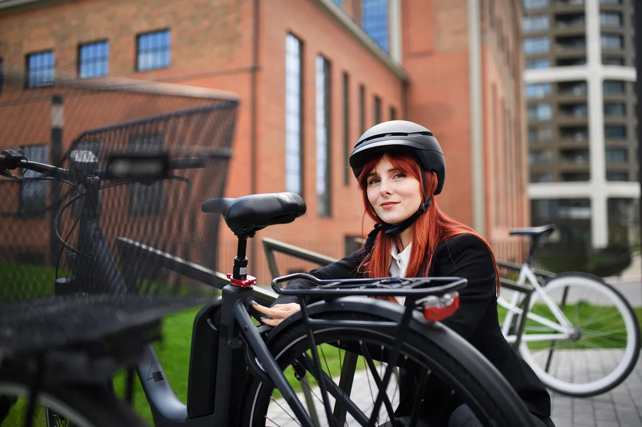 Eine junge Frau mit roten Haaren lächelt in die Kamera, während sie neben einem schwarzen E-Bike steht und einen Helm trägt. Im Hintergrund sind Gebäude zu sehen.