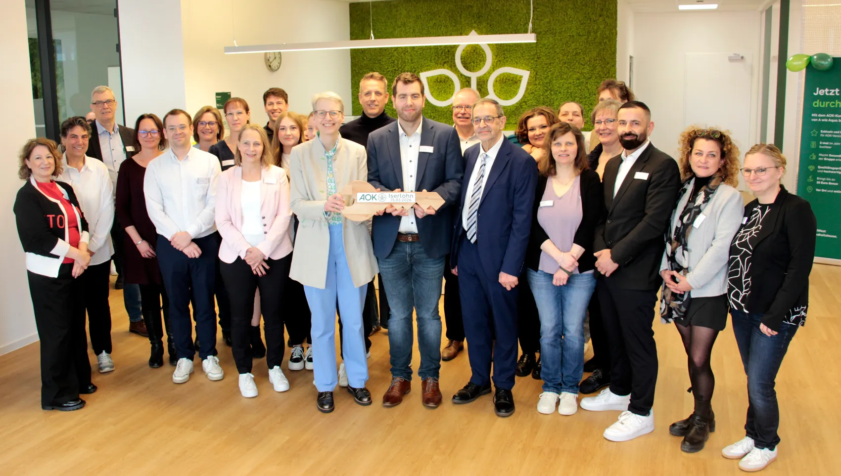 Gruppe von etwa 20 Personen steht lächelnd vor einer grünen Pflanzenwand mit dem AOK-Logo. Im Vordergrund halten zwei Männer ein Eröffnungsband mit dem AOK-Schriftzug.