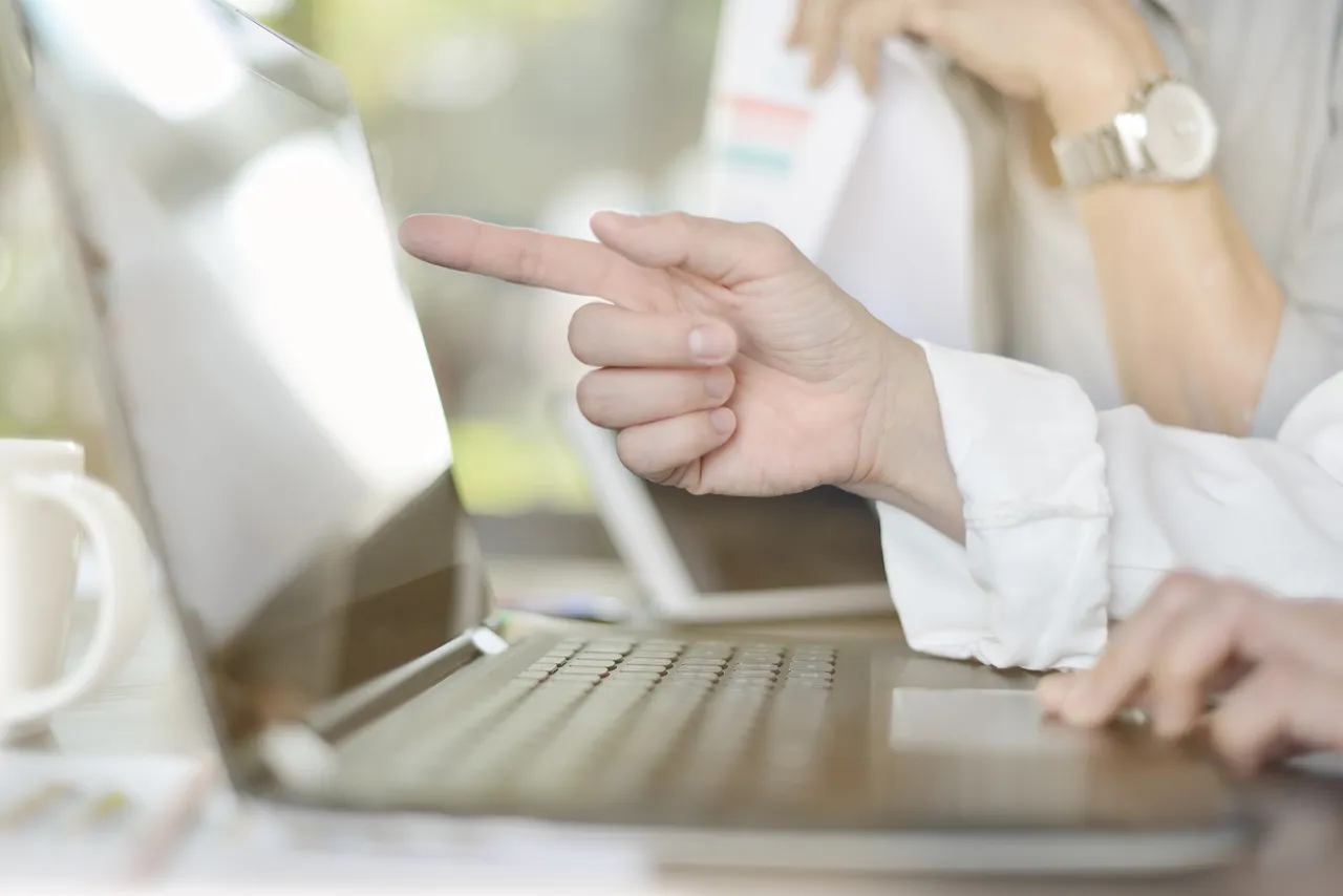 Eine Person zeigt mit dem Finger auf den Bildschirm eines geöffneten Laptops. Im Hintergrund ist eine weitere Hand mit einer Armbanduhr zu sehen.
