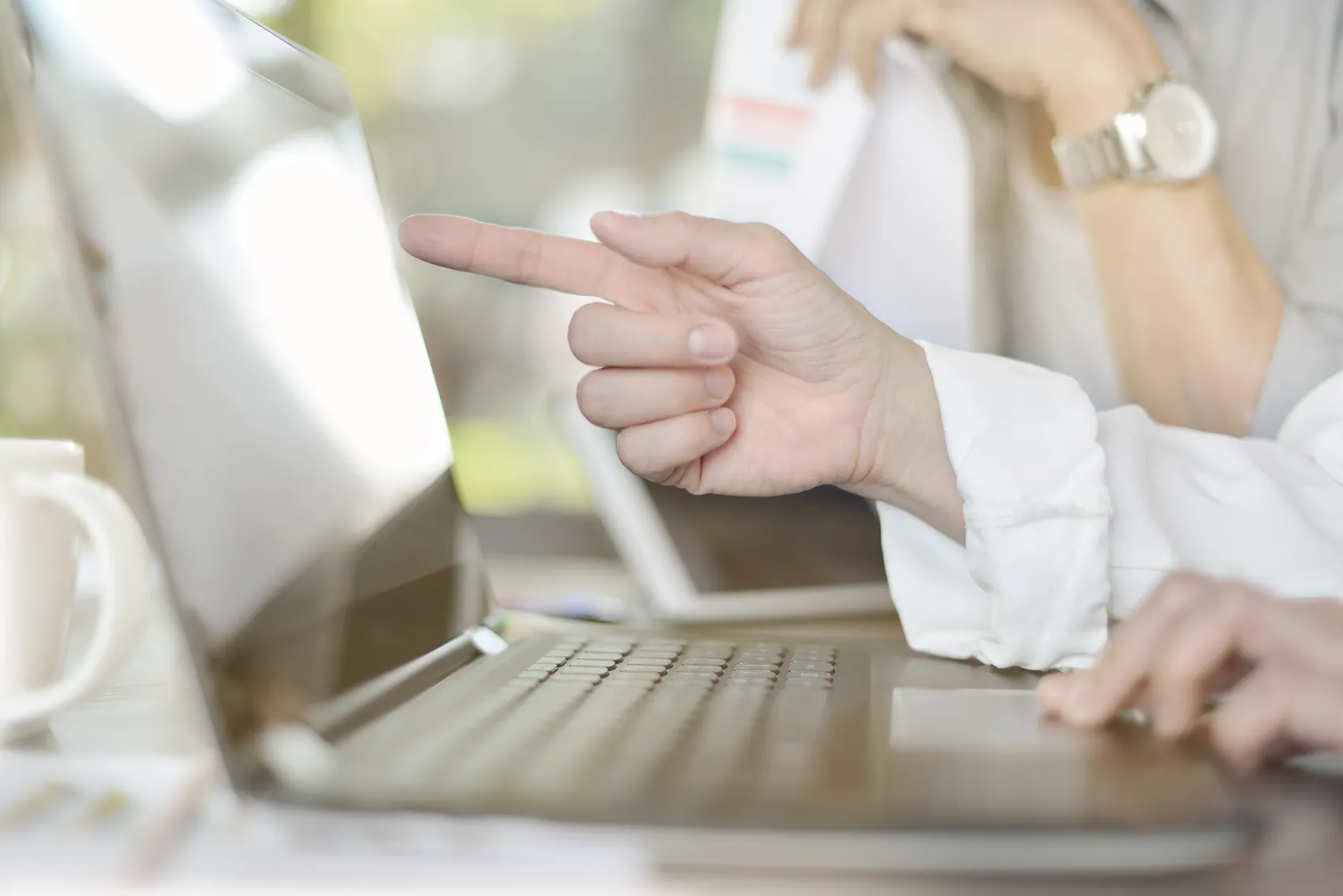 Eine Person zeigt mit dem Finger auf den Bildschirm eines geöffneten Laptops. Im Hintergrund ist eine weitere Hand mit einer Armbanduhr zu sehen.
