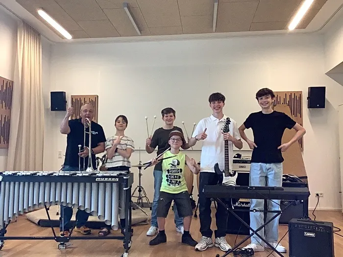 Sechs junge Musiker stehen in einem Proberaum vor ihren Instrumenten: ein Mann spielt ein Xylophon, ein Jugendlicher eine Trompete, ein weiterer eine E-Gitarre, ein anderer ein Keyboard und zwei weitere stehen daneben.