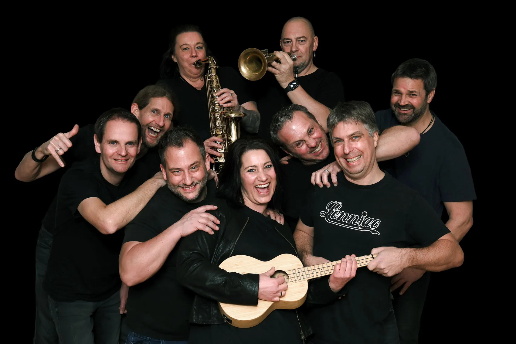 Gruppe von acht lächelnden Musiker*innen, einige mit Instrumenten wie Saxophon und Trompete, posieren eng zusammen. Eine Frau spielt Ukulele im Vordergrund.