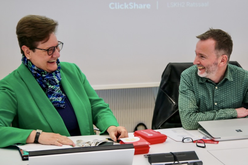 Zwei lächelnde Personen, Kreisdirektorin Barbara Dienstel-Kümper links und Ausschussvorsitzender Denis Potschin rechts, sitzen an einem Tisch und blicken auf Dokumente.