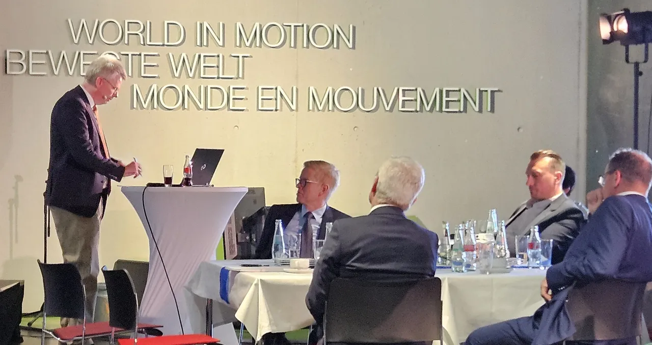 Hubertus Bardt spricht vor einem Tisch mit vier Männern, im Hintergrund ein Wandbild mit dem Schriftzug „World in Motion“ in drei Sprachen.