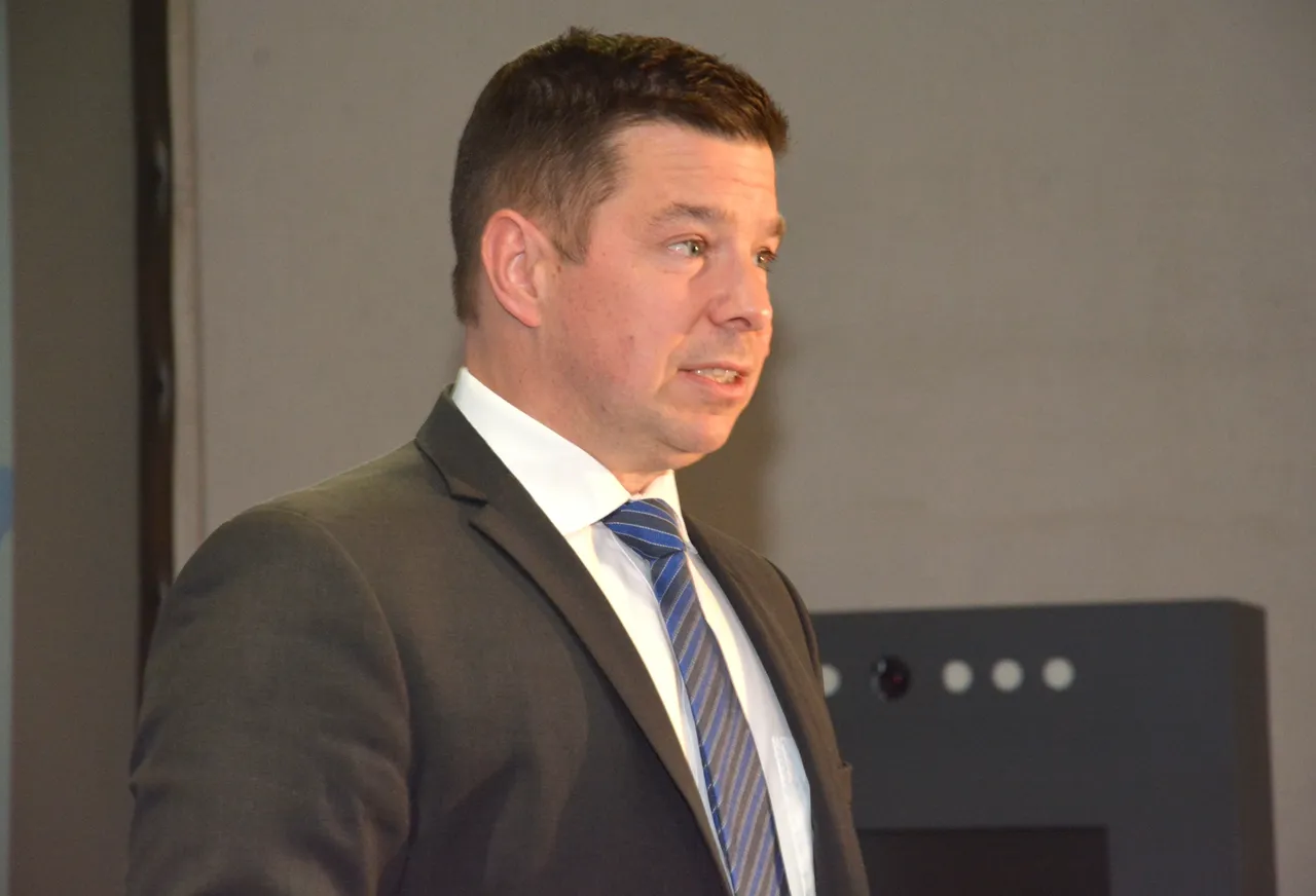 Andreas Kostal spricht auf einer Veranstaltung. Er trägt einen dunklen Anzug, ein helles Hemd und eine blau gestreifte Krawatte.