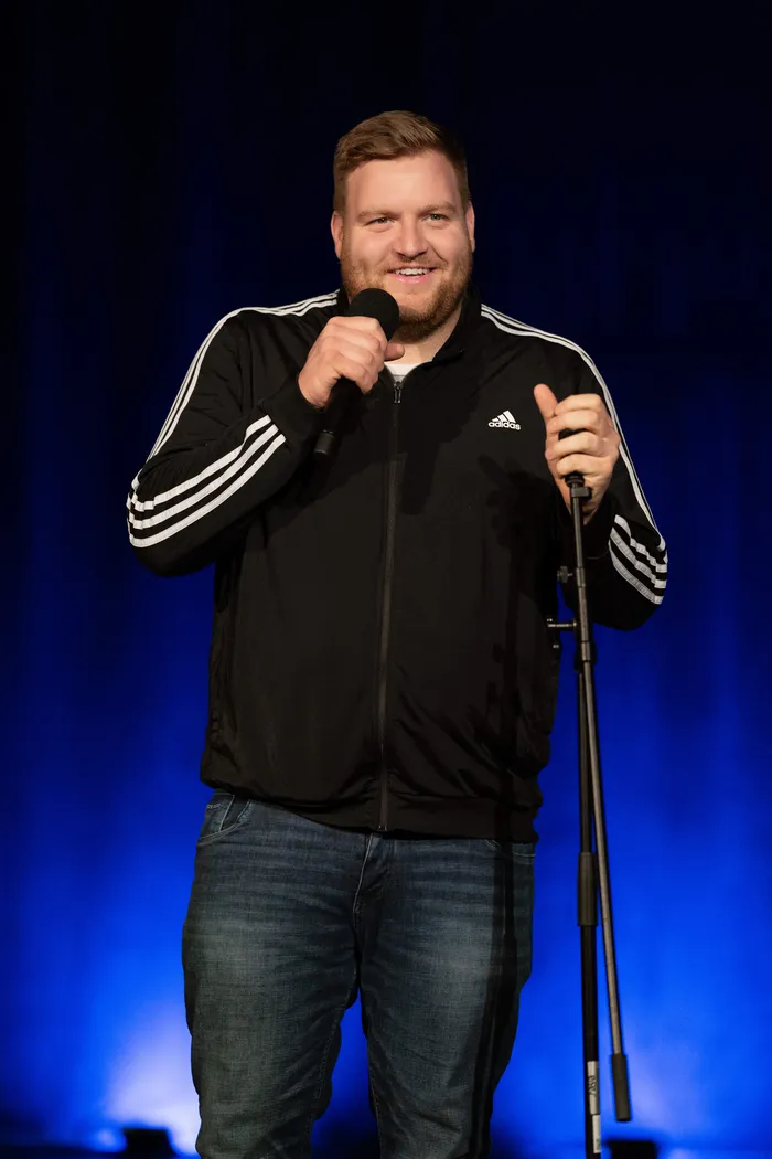 Tobias Beitzel lächelt auf einer Bühne und hält ein Mikrofon sowie einen Mikrofonständer. Er trägt eine schwarze Adidas-Jacke und Jeans.