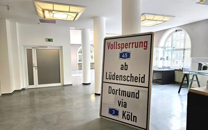 Schild kündigt Vollsperrung der A45 ab Lüdenscheid über Dortmund via Köln an. Im Hintergrund ein heller Flur mit Türen und Schaltern.