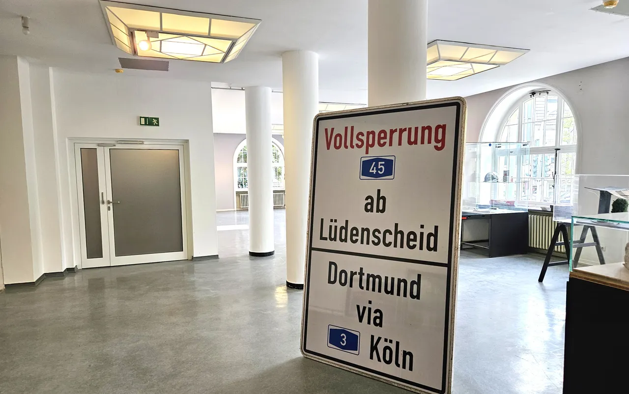 Straßenschild kündigt eine Vollsperrung auf der A45 ab Lüdenscheid über Dortmund via Köln an. Im Hintergrund ist ein heller Flur mit Fenstern und Büromöbeln zu sehen.