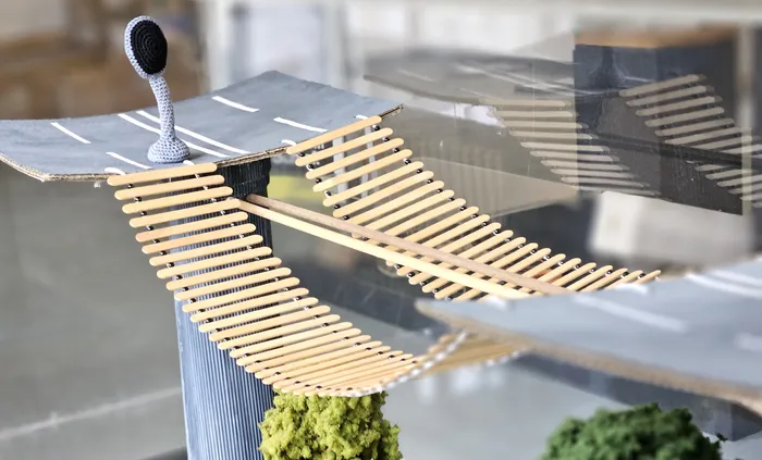 Modell einer Brücke überspannt eine Straße, mit einer Windmühle auf der einen Seite. Die Brücke ist aus Holzlatten gebaut und steht auf einem Fundament aus grünem Moos.