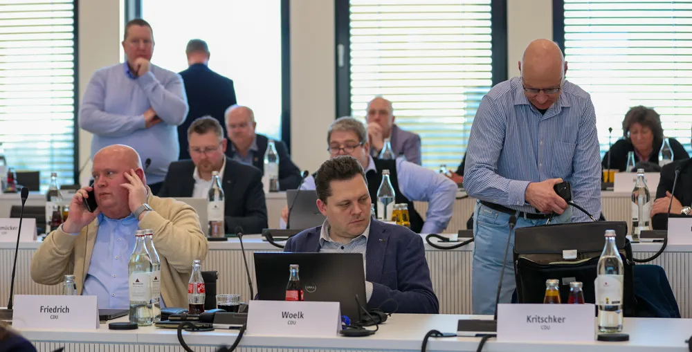 Mehrere Männer und wenige Frauen sitzen an einem Konferenztisch, einige mit Laptop, andere telefonieren oder schauen zu. Namensschilder vor ihnen zeigen Friedrich, Woelk und Kritschker mit dem Zusatz CDU.