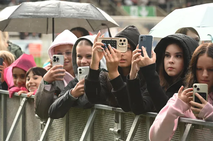 Eine Gruppe junger Menschen filmt mit ihren Smartphones ein Konzert hinter einer Metallabsperrung bei Regen.