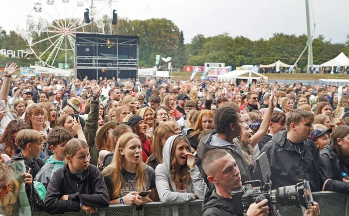 Eine große Menschenmenge bei einem Open-Air-Festival vor einer Bühne, im Hintergrund ist ein Riesenrad zu sehen.