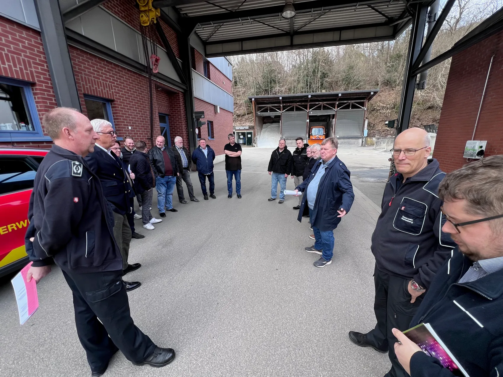 Eine Gruppe von etwa 15 Männern steht vor einer Fahrzeughalle der Feuerwehr und bespricht etwas. Im Vordergrund stehen zwei Männer in Uniform, einer hält Papiere in der Hand.