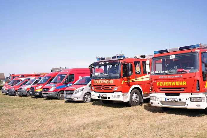 Eine Reihe verschiedener roter Feuerwehrfahrzeuge und Einsatzwagen steht auf einer Wiese unter blauem Himmel.