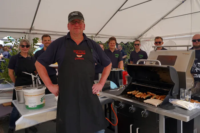 Ein Mann in einer schwarzen Schürze mit der Aufschrift „Bergfest Mannschaft Gelbteam“ steht vor einem Grill unter einem weißen Festzelt.