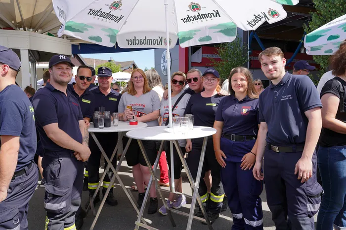 Eine Gruppe von Menschen in dunkelblauen Uniformen und Freizeitkleidung posiert lächelnd um Stehtische unter weißen Sonnenschirmen.