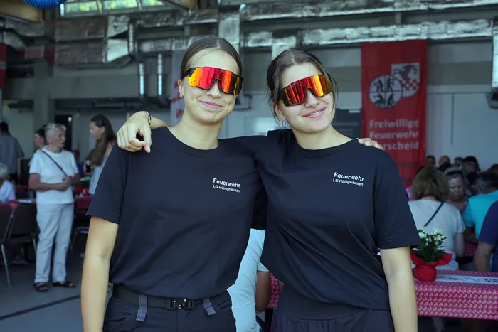 Zwei lächelnde Frauen in schwarzen T-Shirts der Feuerwehr LG Hünghausen tragen große, orange verspiegelte Sonnenbrillen.
