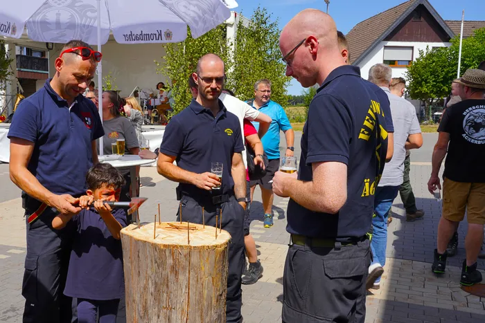 Ein Mann hilft einem Jungen, Nägel in einen Holzstamm zu schlagen, während andere Personen bei einem Fest im Freien daneben stehen.