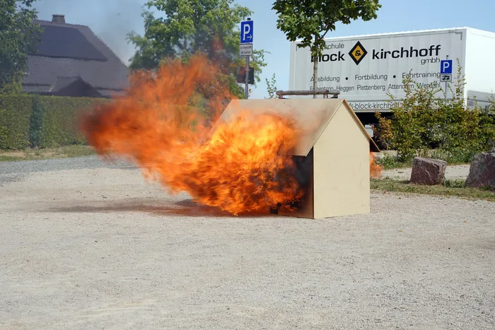 Große Flammen schlagen aus einer kleinen Holzkonstruktion auf einem Schotterplatz hervor.