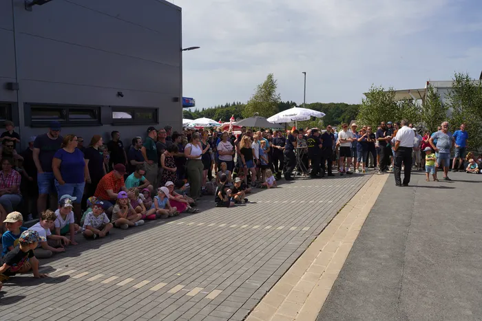 Eine Menschenmenge und sitzende Kinder versammeln sich auf einem gepflasterten Platz vor einem grauen Gebäude beim Bergfest der Löschgruppe Rärin.