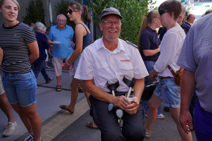 Ein lächelnder Feuerwehrmann in weißer Uniform mit der Aufschrift „FEUERWEHR“ hält ein Glas Bier beim Bergfest der Löschgruppe Rärin.