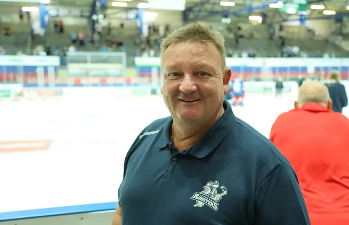 Bernd Schutzeigel lächelt in einem dunkelblauen Polo-Shirt mit Roosters-Logo vor dem Hintergrund einer Eishalle.
