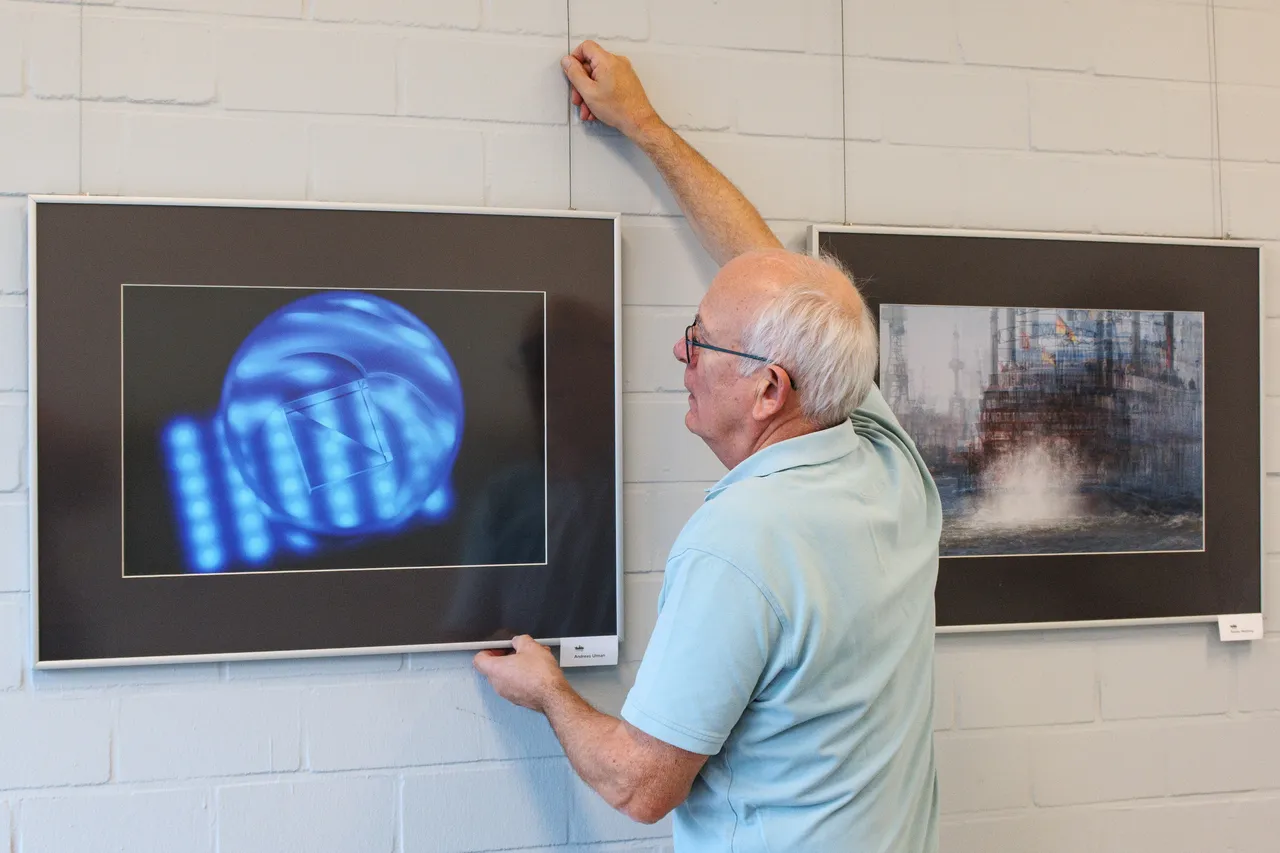 Ein Mann hängt ein gerahmtes Foto an einer weißen Ziegelwand auf, neben einem bereits aufgehängten Bild einer blauen Installation.