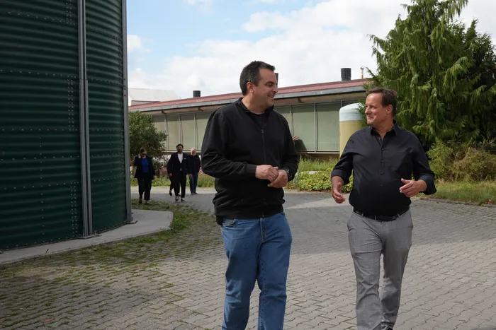 Stephan Werthmann und Thorsten Schick im Gespräch beim Gehen auf einem gepflasterten Weg vor einem großen grünen Silo.