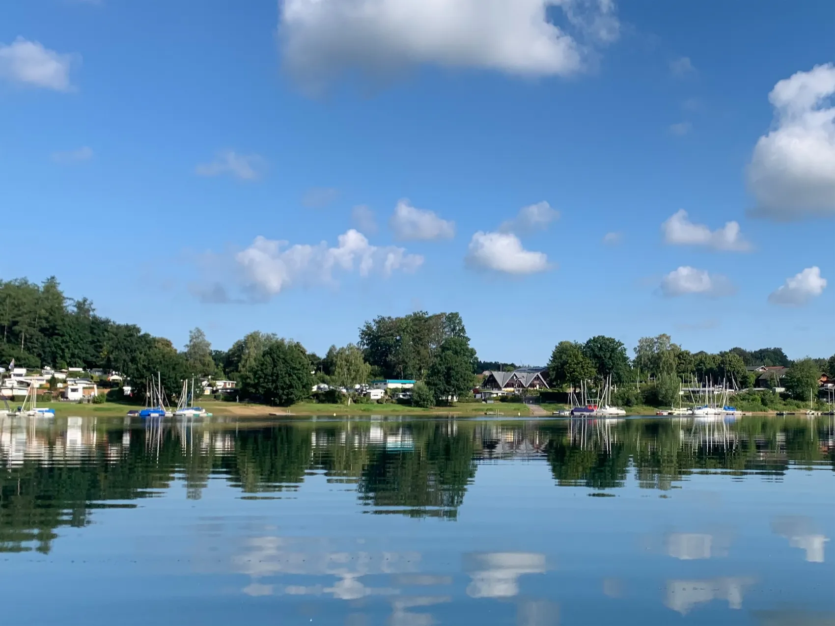 Die Bever-Talsperre liegt ruhig im Sonnenschein, umgeben von Bäumen und Häusern. Mehrere Segelboote treiben auf dem Wasser, das den blauen Himmel und die Wolken spiegelt.