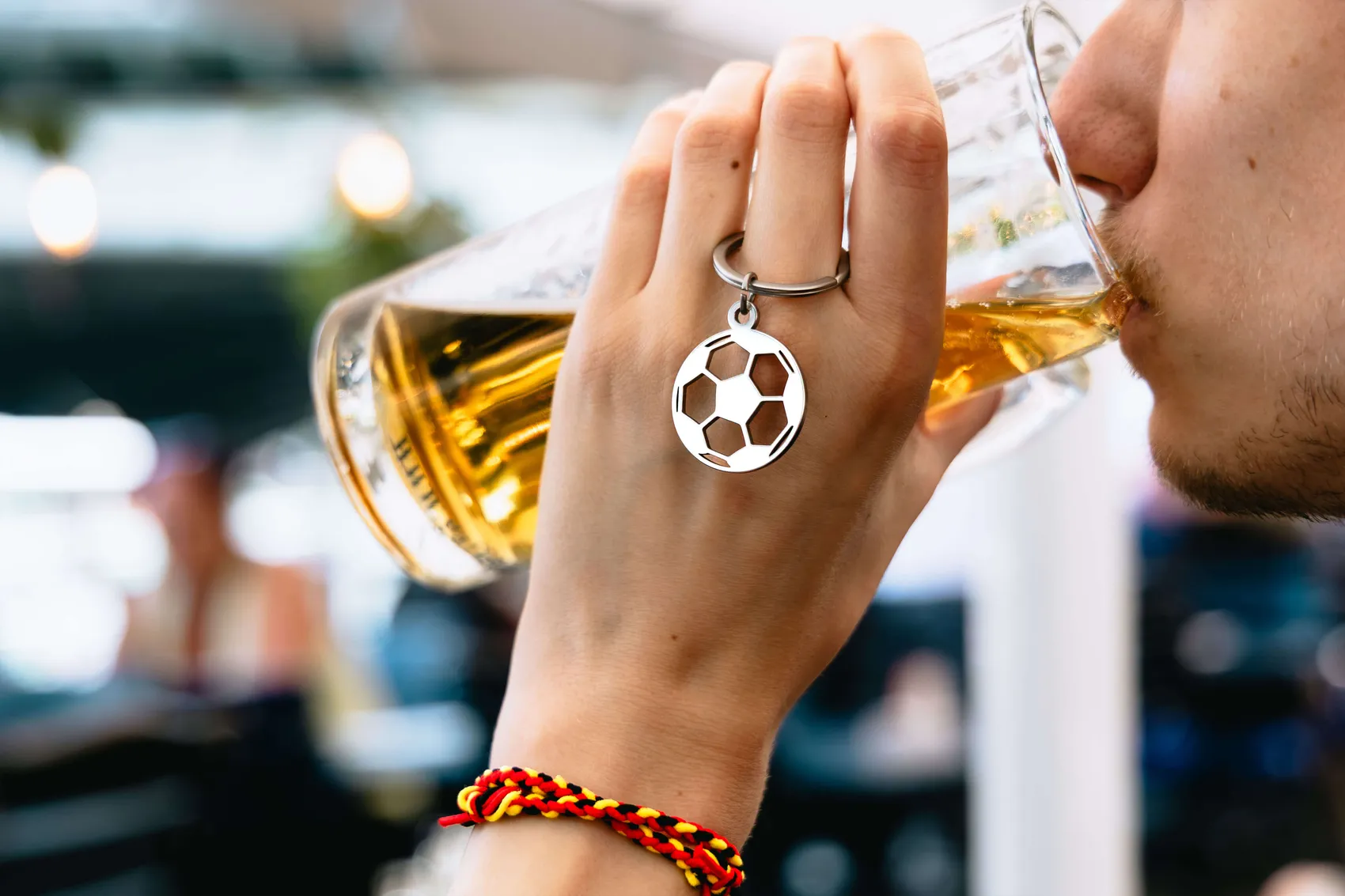 Eine Hand mit einem Fußball-Schlüsselanhänger und einem roten Armband hält ein Bierglas an. Im Hintergrund ist ein Mann mit Bart zu sehen, der aus dem Glas trinkt.