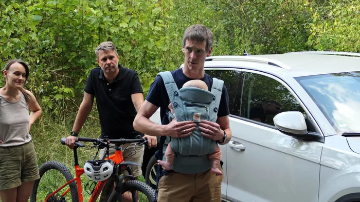 Ein Mann trägt ein Baby in einer Tragehilfe, während im Hintergrund ein Mann bei einem Mountainbike und eine Frau stehen.