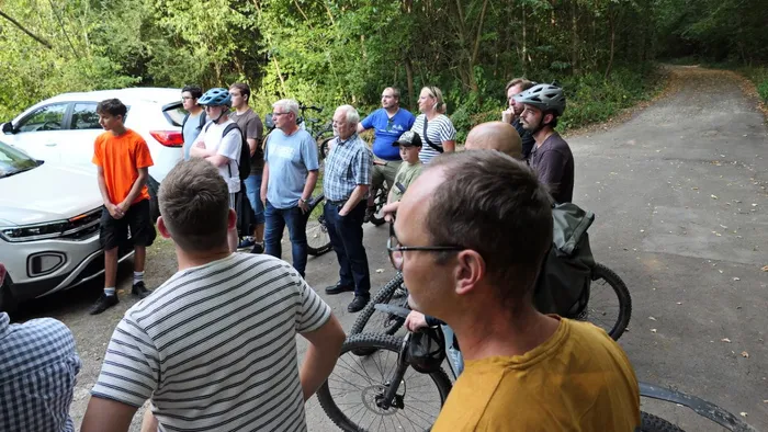 Eine Gruppe von Menschen steht auf einem asphaltierten Weg in einem Waldgebiet neben geparkten Autos und Fahrrädern.