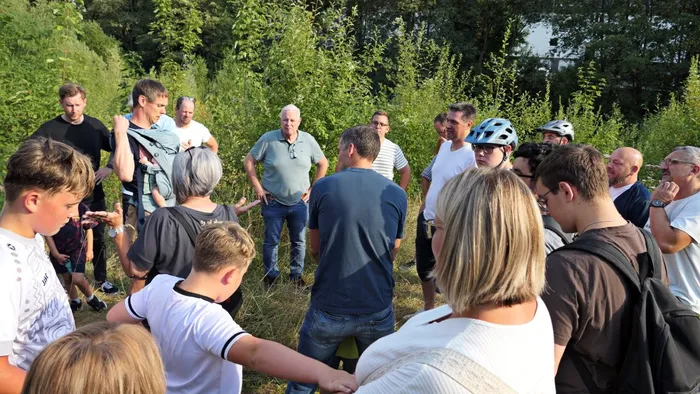 Eine Gruppe von Menschen steht im Gespräch zusammen in einer Grünanlage mit Büschen und Bäumen im Hintergrund.