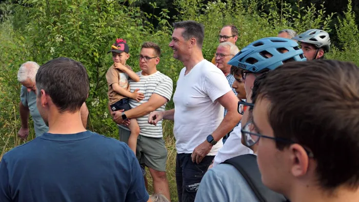 Eine Gruppe von Menschen, teils mit Fahrradhelmen, steht bei einem Treffen im Freien vor grüner Vegetation.