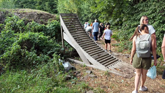 Eine Gruppe von Menschen steht vor einer großen Holzrampe in einem bewaldeten Gelände.