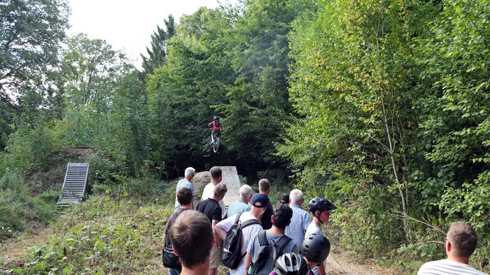 Ein Mountainbiker springt in der Luft, während eine Gruppe von Menschen in einem bewaldeten Bikepark zuschaut.