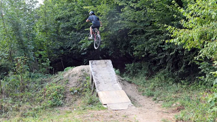 Ein Mountainbiker springt von einer Holzrampe auf einem Erdhügel in einem Waldgebiet ab.