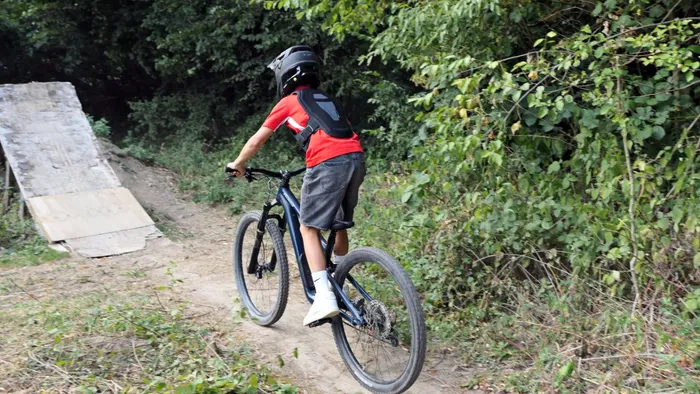 Ein Mountainbiker mit Helm und rotem T-Shirt fährt auf einem Pfad in einem Bikepark auf eine Holzrampe zu.