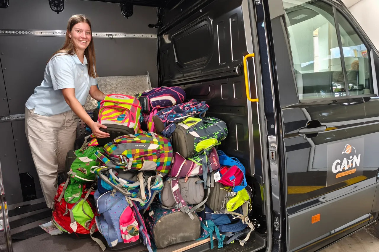 Eine junge Frau steht neben einem Transporter, der mit vielen bunten Schultornistern beladen ist.